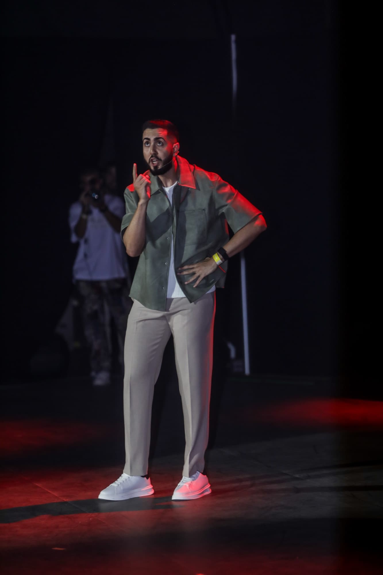 Elías in a sage jacket pointing to the crowd under red rim light