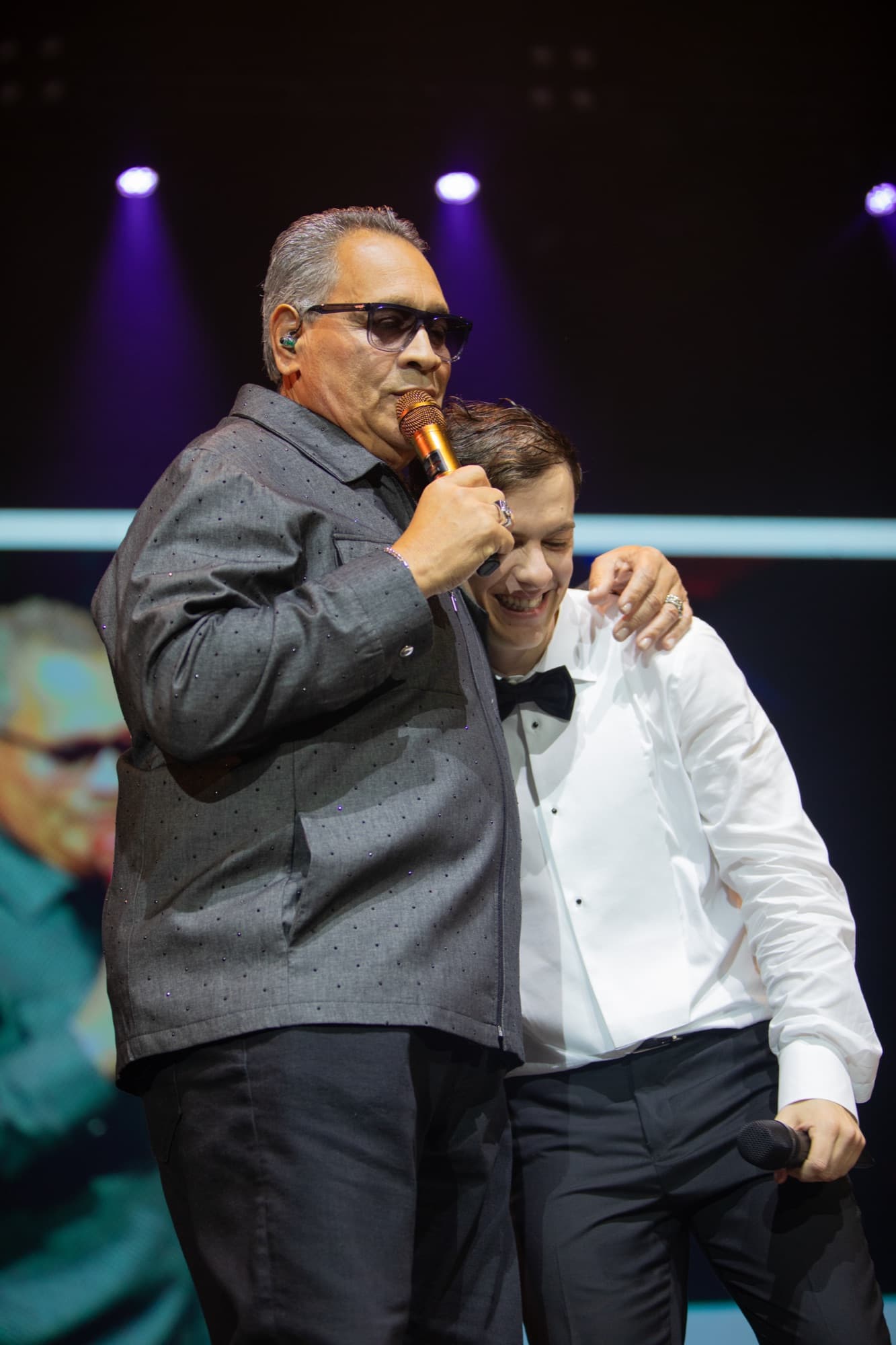 Veteran singer at the mic with his arm around a young guest in a tuxedo
