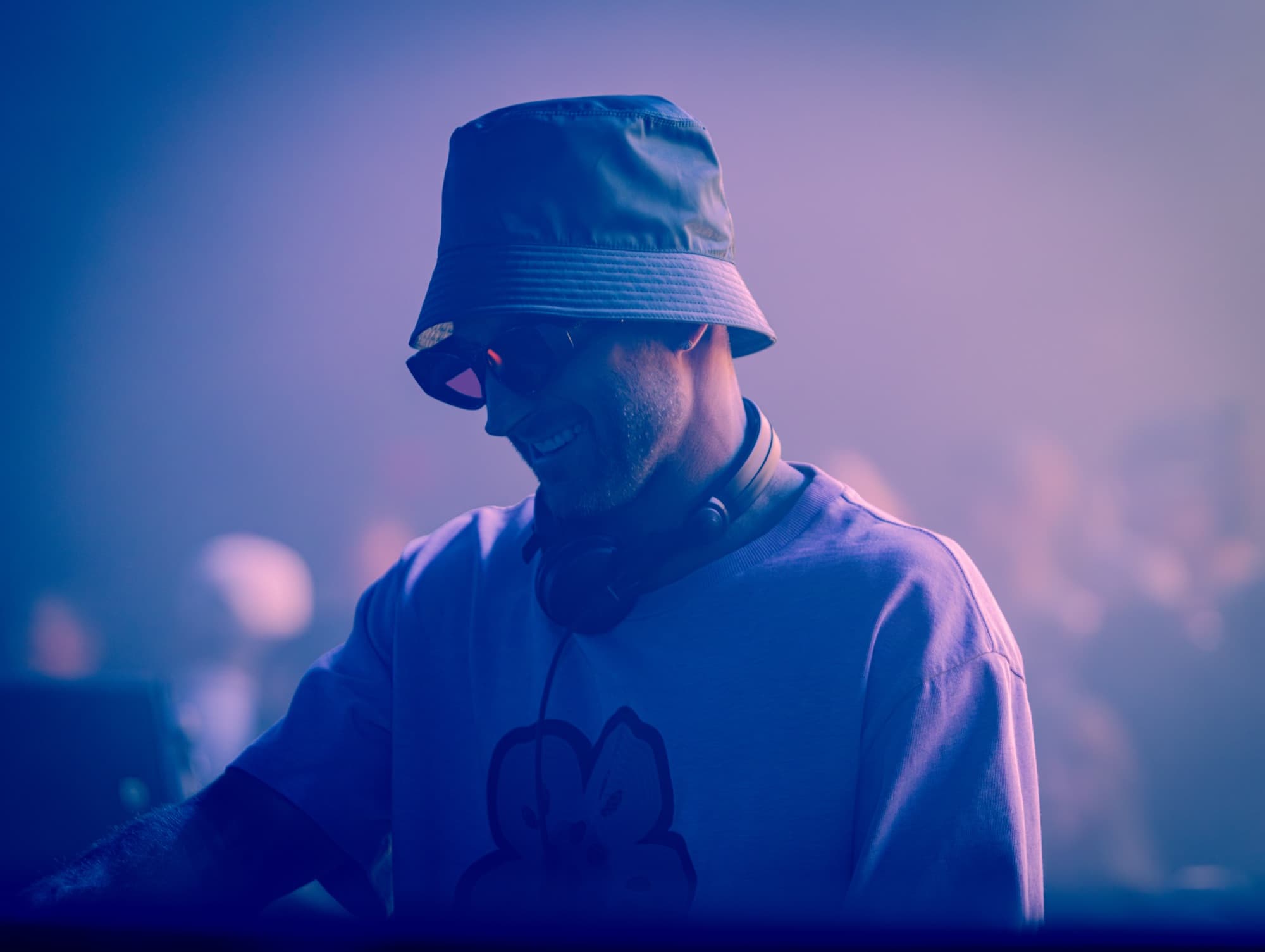 DJ in a bucket hat smiling through violet haze mid-set