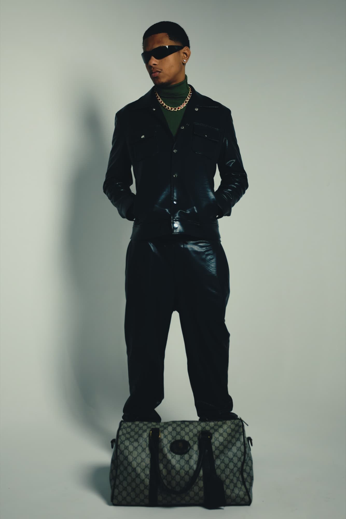 Model in black patent leather with a monogram duffel at his feet