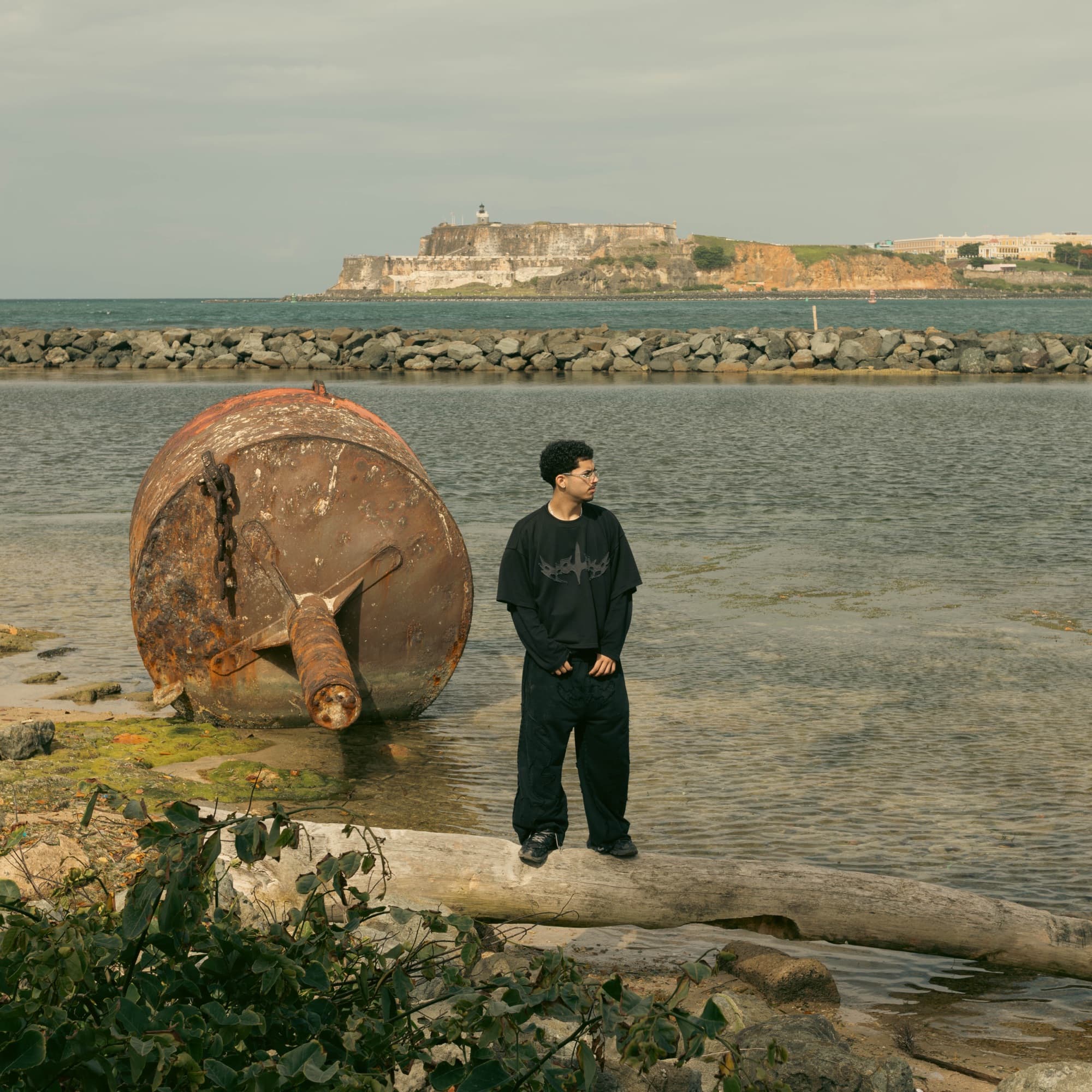 Model standing on driftwood with El Morro across the bay